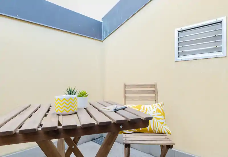 Pequeña terraza privada con mesa y silla de madera, cojín decorativo y suculentas en maceta — espacio exterior luminoso y bien arreglado.