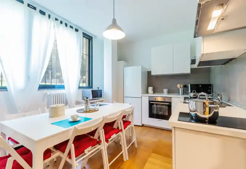 Cocina moderna y luminosa con mesa para seis, muebles blancos y una gran ventana que aporta luz natural — ideal para comidas familiares.