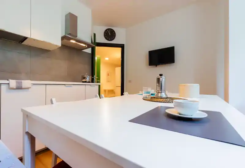 Cocina moderna y luminosa con una gran mesa blanca puesta con una taza, un mantel individual y una cafetera moka; armarios limpios y zona de cocina compacta al fondo.