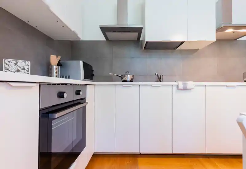 Cocina moderna y luminosa con muebles blancos, electrodomésticos de acero inoxidable y protector de pared de azulejos limpio. La imagen destaca el horno empotrado, la placa y el fregadero — perfecta para anuncios de alquiler.