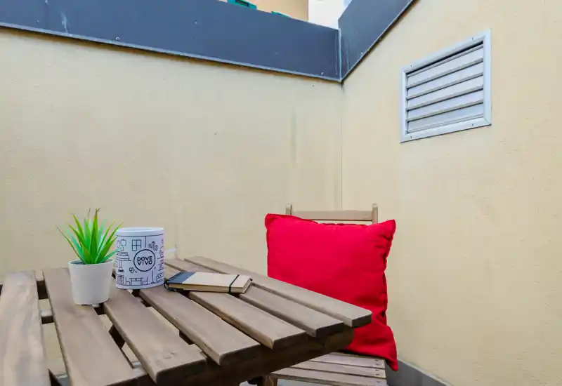 Pequeño balcón privado con mesa y silla de madera, cojín rojo y planta en maceta — ideal para tomar un café o leer.