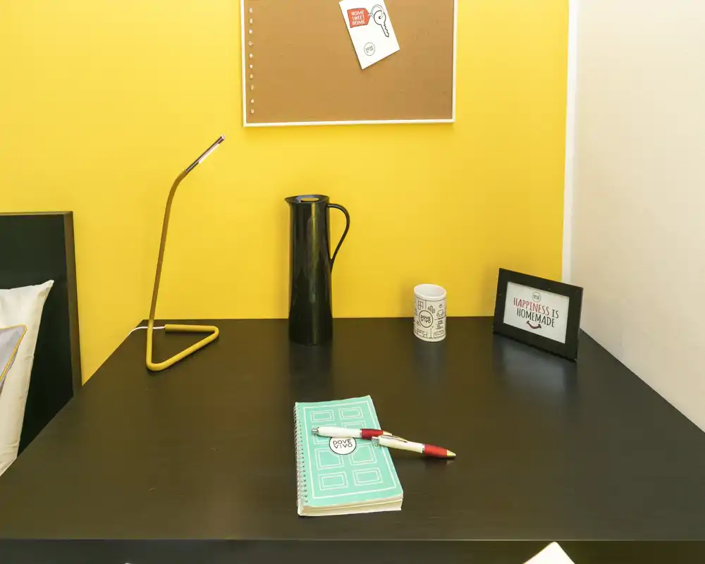 Coin de chambre compact avec un bureau foncé contre un mur jaune vif, présentant une lampe, des objets décoratifs et une tête de lit partiellement visible.