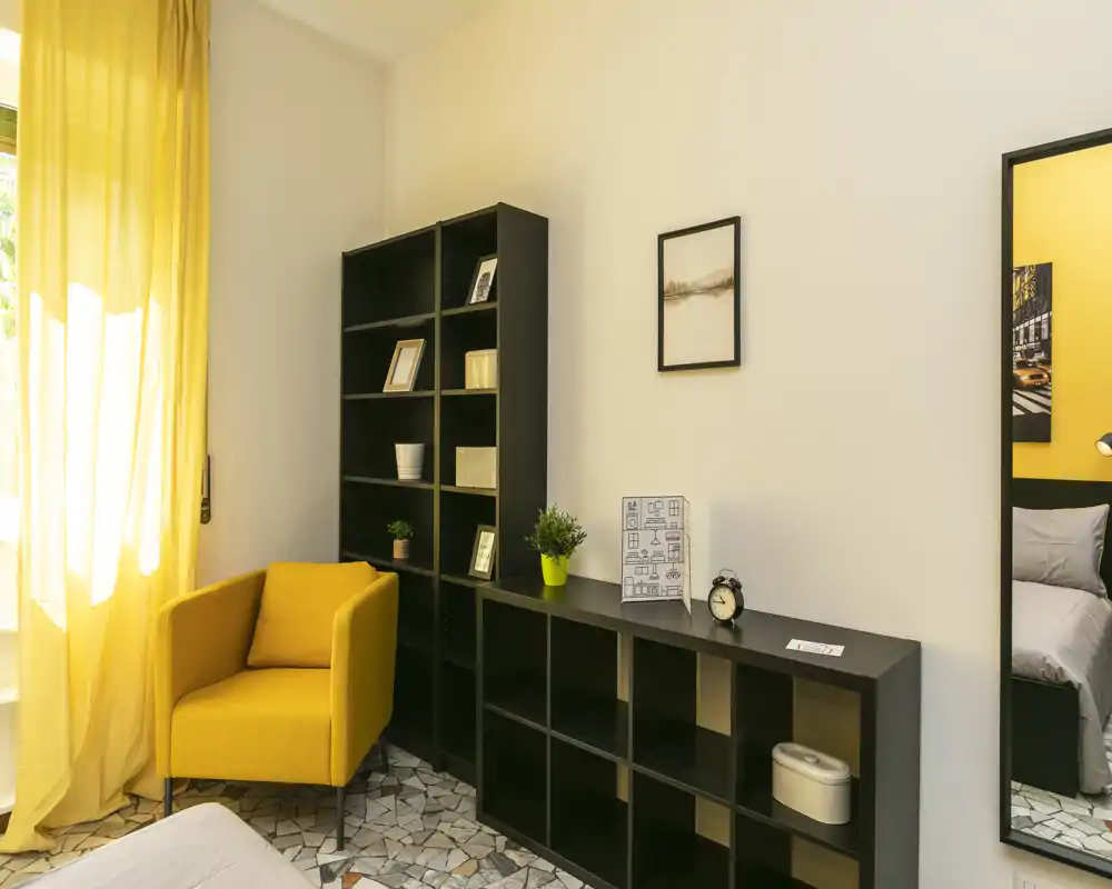 Coin de chambre lumineux et chaleureux avec un fauteuil moutarde, des étagères et un lit partiellement visible ; des touches jaunes et la lumière naturelle donnent une ambiance accueillante.