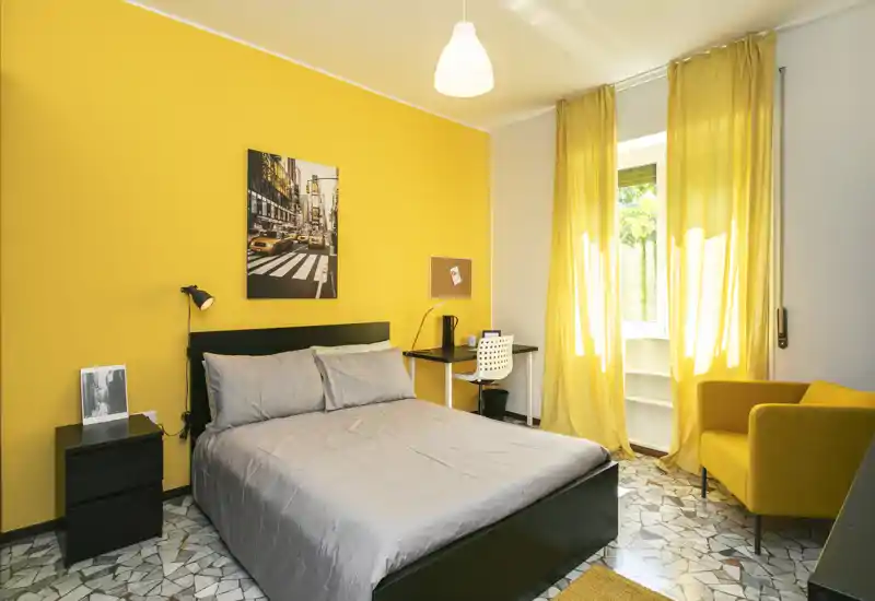 Chambre lumineuse avec grand lit, accents jaunes assortis et un petit bureau près de la fenêtre. Décor moderne et chaleureux avec un fauteuil et un sol en carreaux.