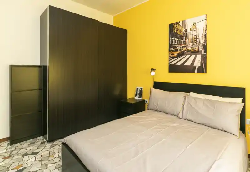 Chambre moderne et lumineuse avec lit double, grand placard en bois foncé et mur jaune chaleureux; linge de lit soigné et table de chevet pour une atmosphère cosy.