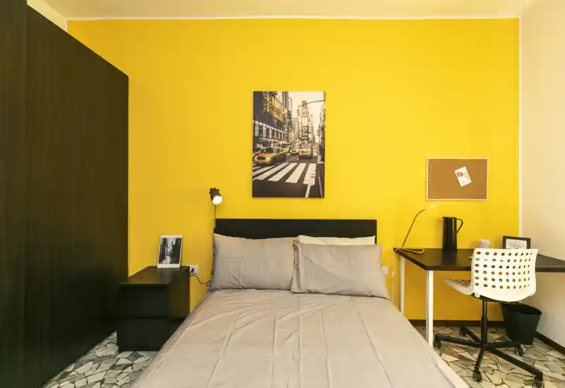 Chambre lumineuse et bien mise en scène avec un lit double devant un mur jaune vif, table de chevet et bureau compact — parfaite pour des séjours confortables.