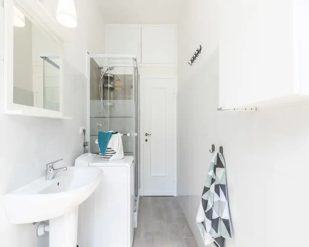 Salle de bains lumineuse et moderne avec lavabo sur pied, cabine de douche en verre et machine à laver ; carrelage blanc et sol imitation bois pour un style frais et minimaliste.