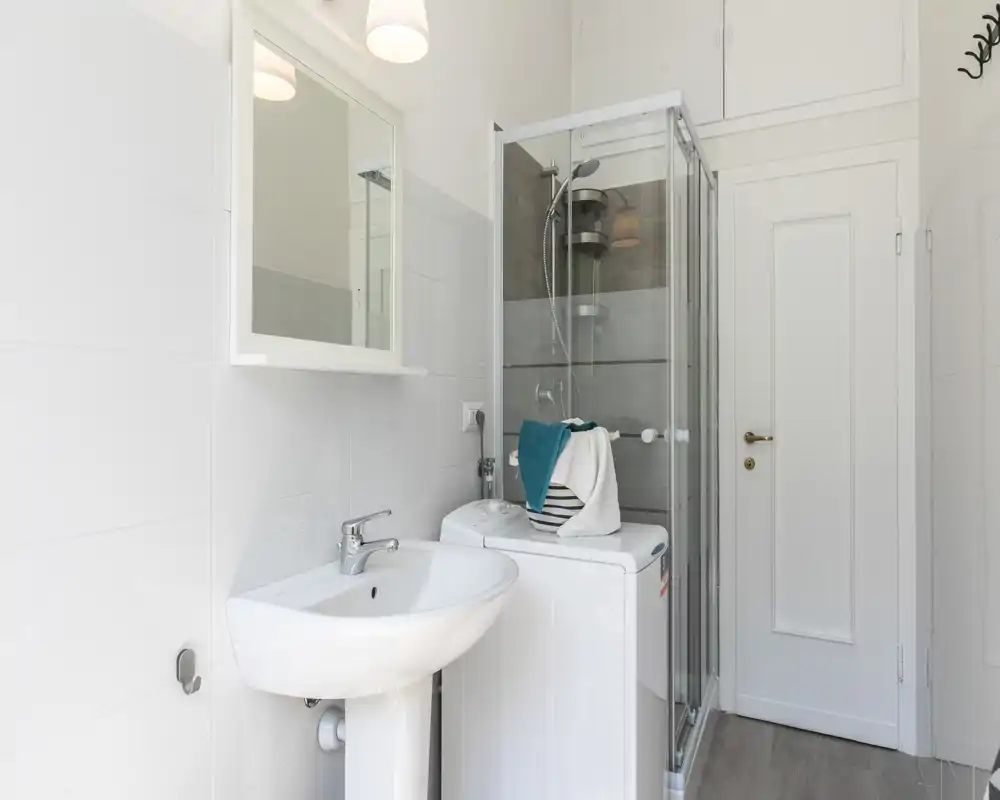 Salle de bains propre et compacte avec lavabo sur pied, armoire miroir, cabine de douche vitrée et machine à laver ; carrelage blanc lumineux et équipements modernes, pratique pour la location.