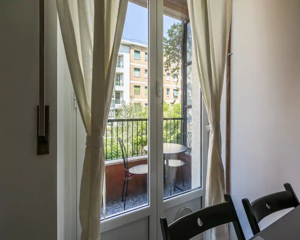 Petit balcon ensoleillé avec une table ronde et deux chaises en métal, visible à travers des baies vitrées et des rideaux légers ; idéal pour le petit-déjeuner face au jardin.