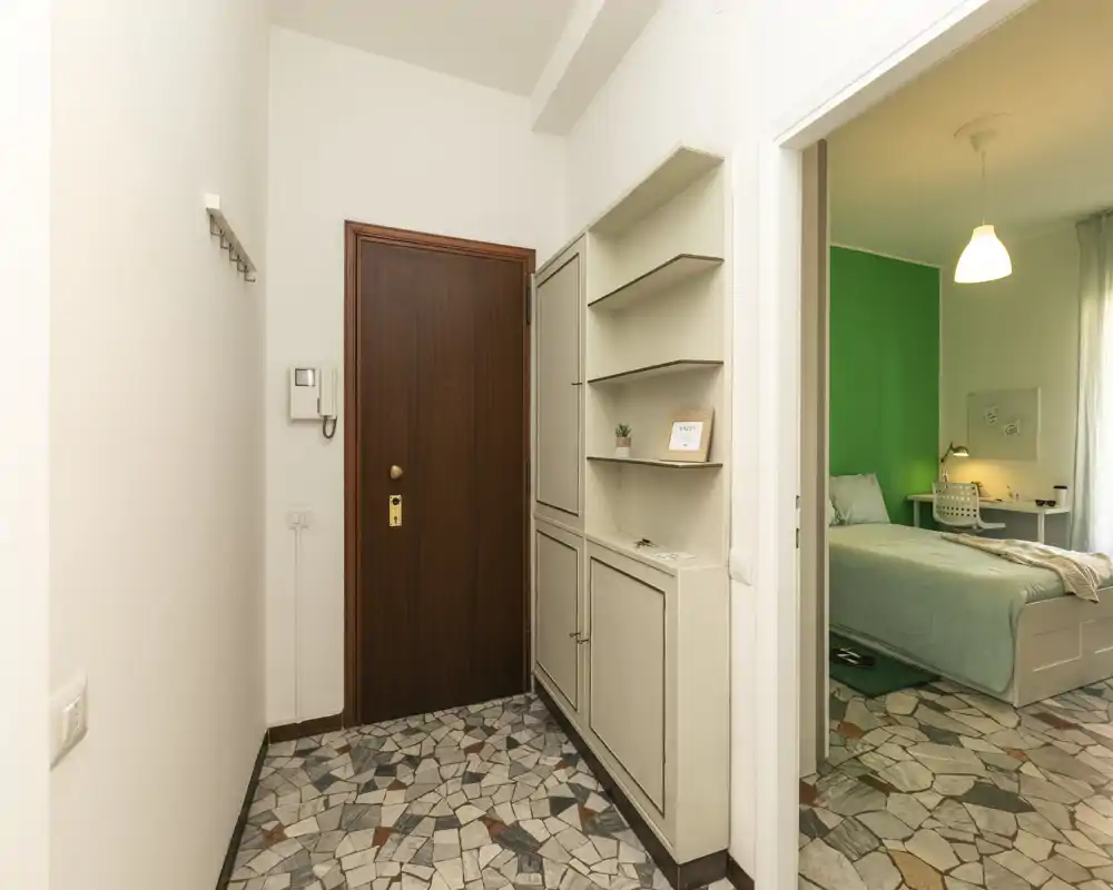 Entrée d'appartement lumineuse et soignée avec une porte d'entrée en bois, un meuble de rangement intégré et un sol en carreaux mosaïque. La chambre adjacente est visible par la porte ouverte.