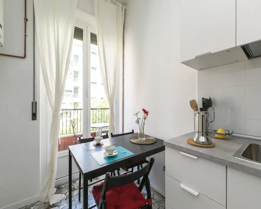 Cuisine compacte et lumineuse avec une petite table pour deux, un évier et un plan de travail avec ustensiles, et une lumière naturelle directe depuis la porte du balcon.