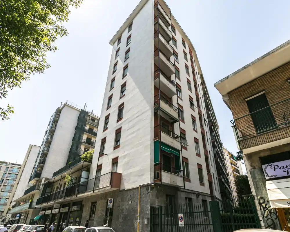 Vue extérieure d'un immeuble résidentiel de plusieurs étages par une journée ensoleillée, avec balcons, commerces au rez-de-chaussée et voitures garées — adapté pour présenter la façade et le quartier.