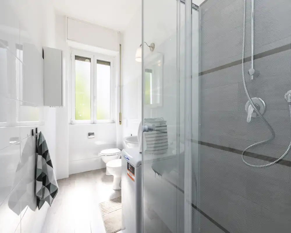 Salle de bain moderne et lumineuse avec cabine de douche en verre, sanitaires muraux, WC et une fenêtre apportant la lumière naturelle.