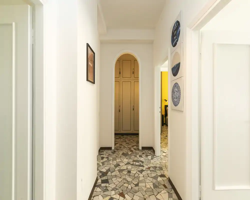 Entrée lumineuse avec sol en mosaïque décorative et une porte en arc menant à un placard intégré. Murs blancs et cadres muraux offrent un espace d'accueil soigné.