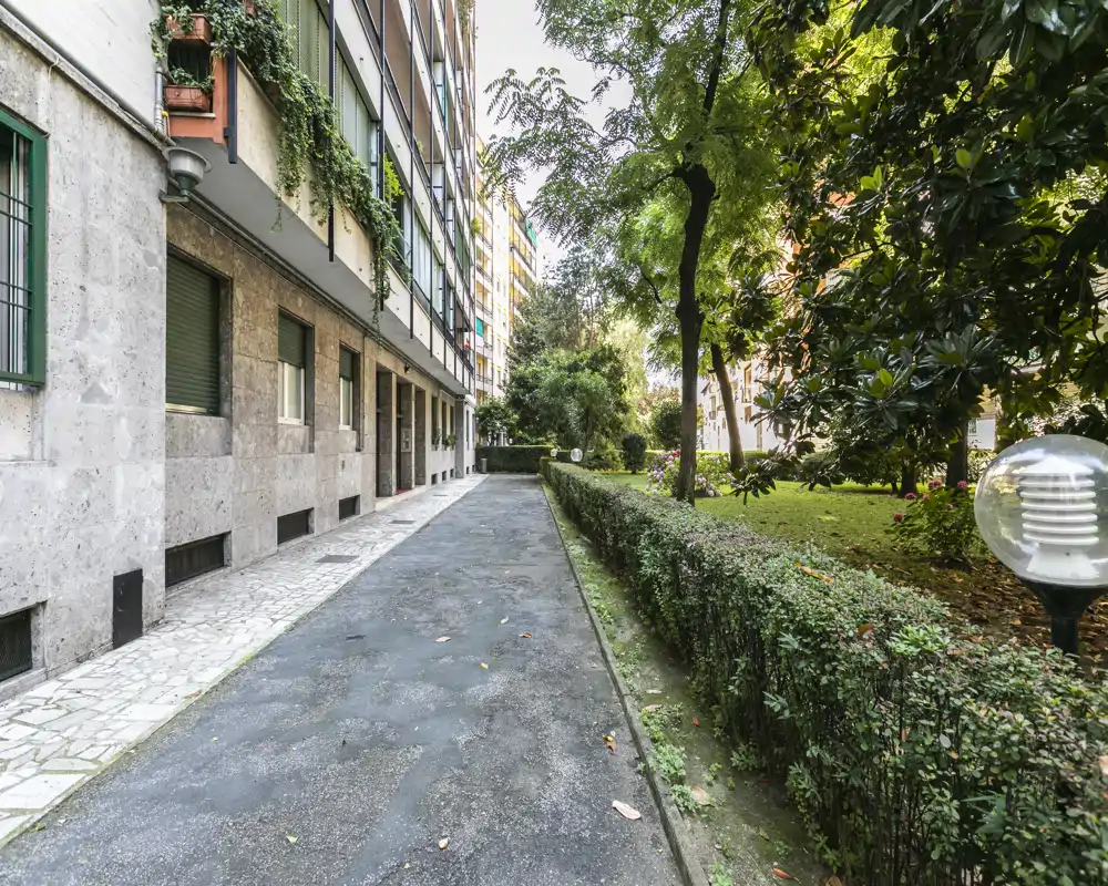 Allée extérieure à côté d'un immeuble résidentiel avec une haie entretenue et un espace vert ; chemin pavé entre l'immeuble et le jardin.