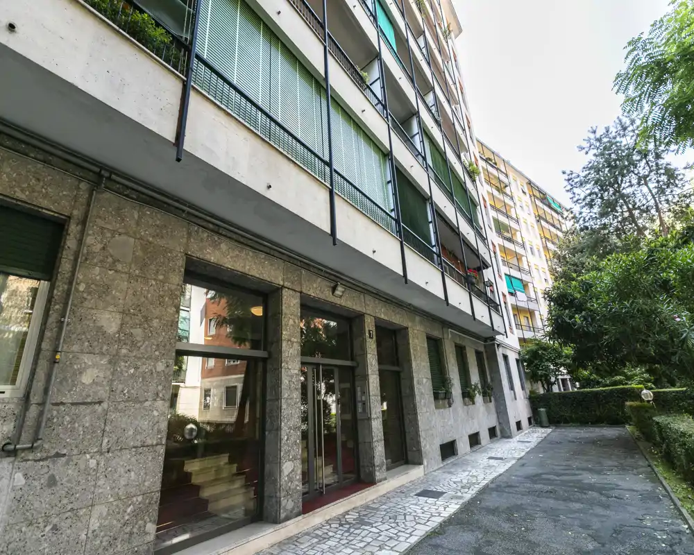 Vue extérieure de l'entrée d'un immeuble résidentiel avec chemin pavé, façade en pierre et espaces verts sur le côté ; idéale pour présenter la façade et l'accès.