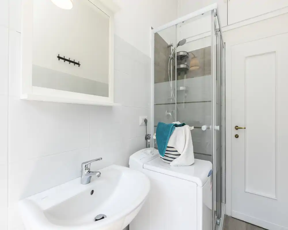 Salle de bain compacte et lumineuse avec lavabo, armoire-miroir, cabine de douche et machine à laver à côté de la douche; carrelage blanc et équipements modernes pour un rendu propre.