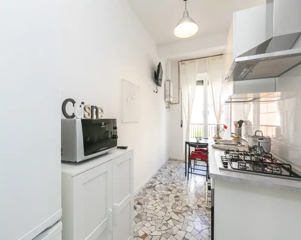 Cuisine lumineuse et compacte avec plaque à gaz, hotte et petit coin repas près d'une fenêtre ensoleillée. Sol en mosaïque et meubles blancs pour une atmosphère propre et accueillante.