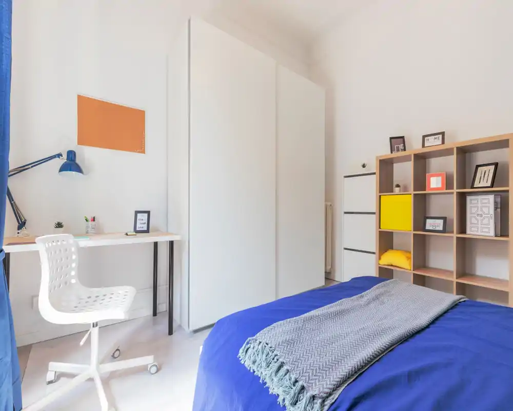 Chambre moderne et lumineuse avec lit habillé de bleu, petit bureau et grand placard blanc; bien éclairée et décorée simplement pour un séjour confortable.