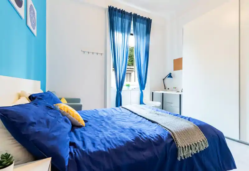 Dormitorio luminoso y bien decorado con cama grande vestida en azul intenso, manta y cojines acogedores, escritorio junto a la ventana y amplio armario. La luz natural y la decoración coordinada generan un ambiente fresco y atractivo.