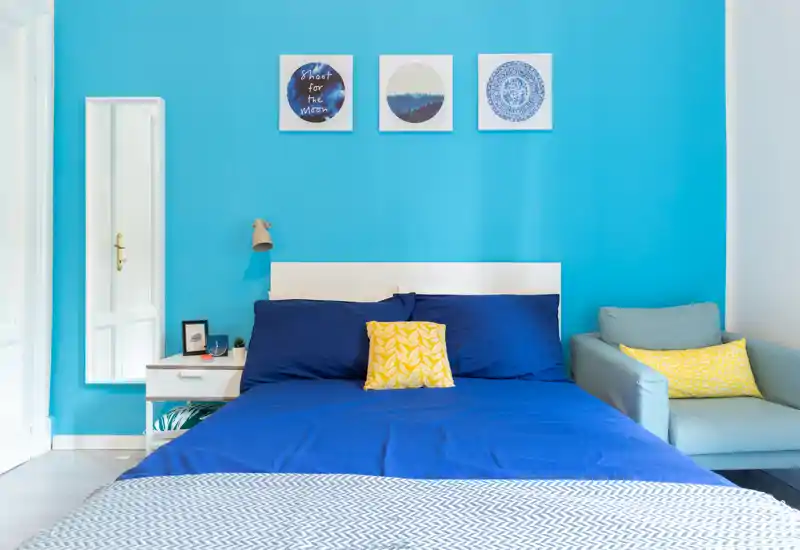 Dormitorio moderno y luminoso con una gran cama azul, cojines a juego y un cómodo sillón frente a una pared turquesa vibrante.