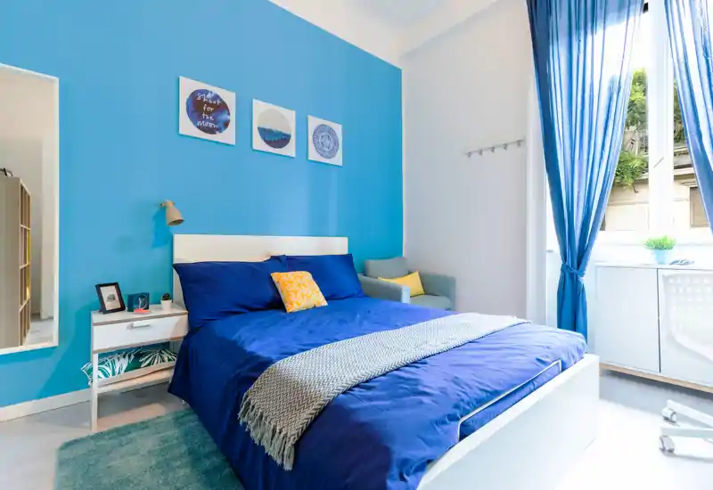 Dormitorio moderno y luminoso con una amplia cama azul, mesita de noche, textiles acogedores y ventana soleada — ideal para destacar el confort y la luz natural.