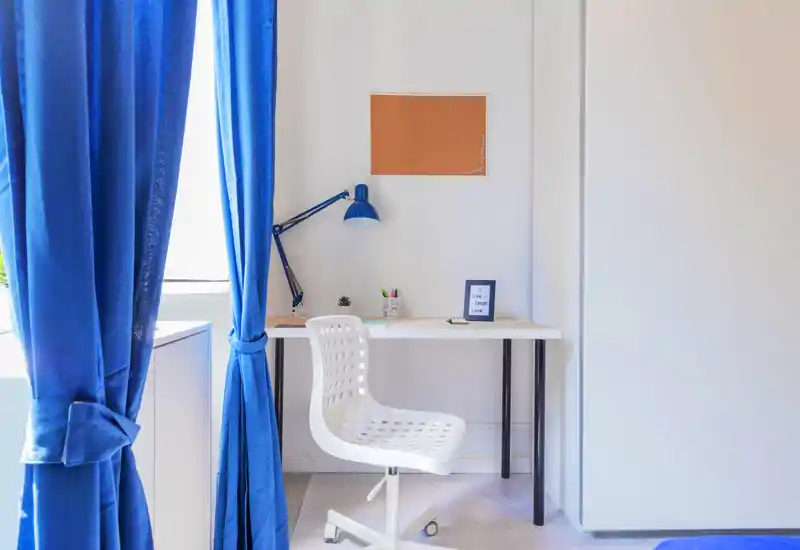 Espacio de trabajo luminoso y minimalista con escritorio blanco, lámpara moderna y una silla giratoria perforada junto a una ventana con cortinas azules.