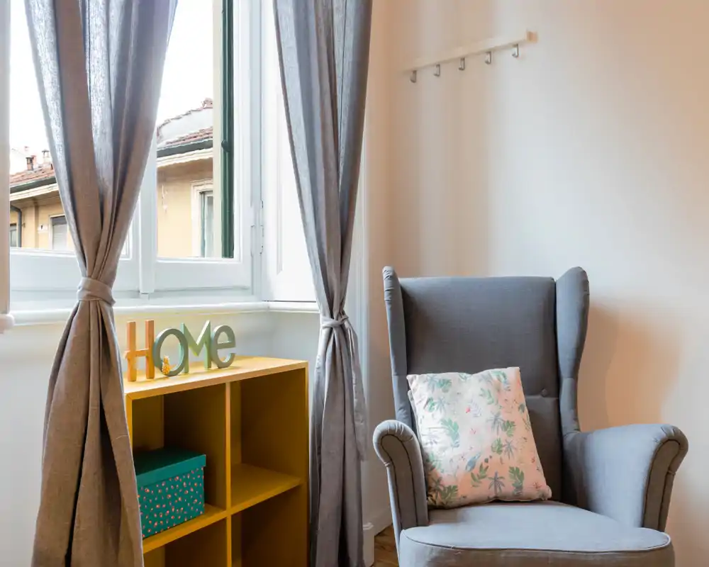 A bright, well-styled living corner featuring a comfortable wingback armchair with a patterned cushion, soft curtains and a small yellow shelving unit with decorative accents.