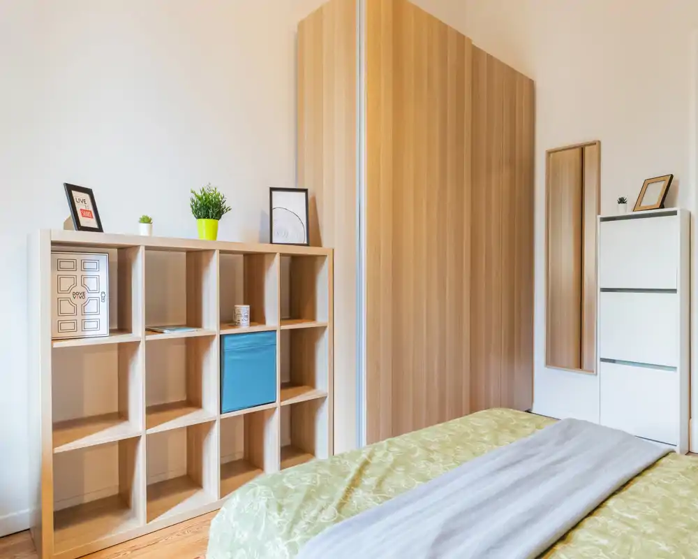 Bright, modern bedroom with a neatly made bed, wooden wardrobe and open shelving providing practical storage; light tones and simple decor create a calm, inviting atmosphere.