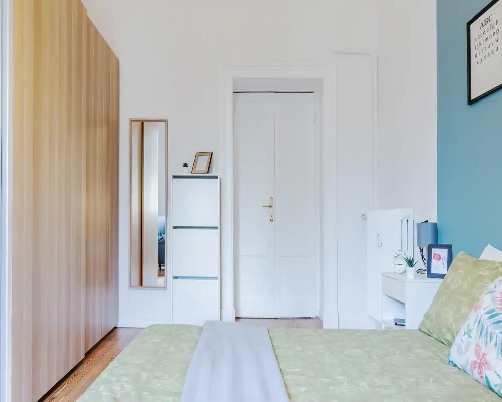 Bright, neatly styled bedroom with a partially visible bed in the foreground, bedside table and wardrobe; clean, airy decor with soft green bedding and tasteful accents.