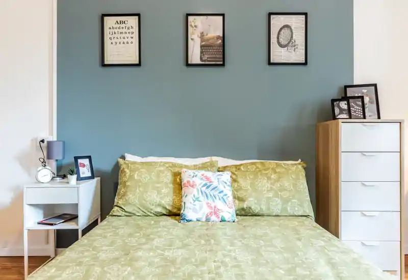 Bright, stylish bedroom with a large bed dressed in green patterned linens, decorative cushions and simple framed artwork above. The room features coordinated bedside furniture and a tall drawer unit for storage.