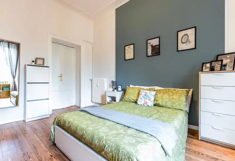 Bright, well-staged bedroom with a double bed dressed in green linens, decorative cushions and framed artwork on a slate-blue accent wall. White storage units and warm wooden floors complete a cozy, modern atmosphere ideal for listings.