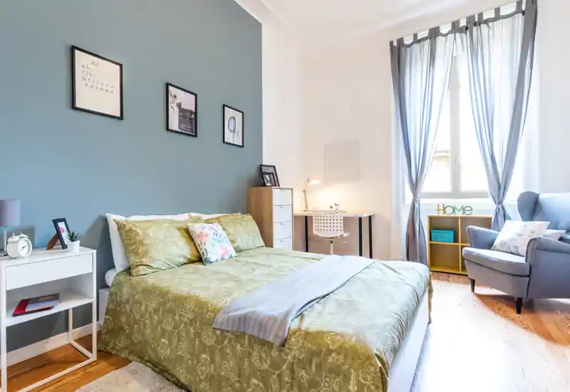 Bright, well-staged double bedroom with a neatly made bed, desk by the window and a comfortable armchair — ideal for a cozy stay with lots of natural light.