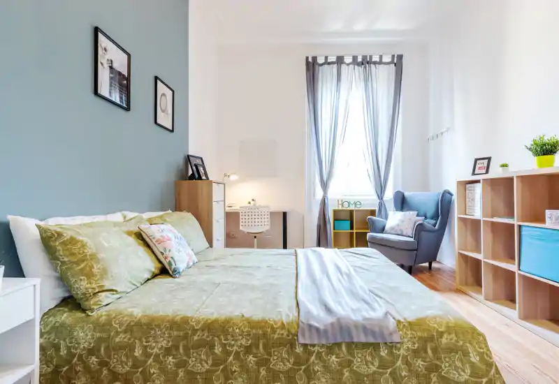 Bright, well-styled bedroom with a large bed in the foreground, a cozy armchair by the window and a small desk — ideal for a comfortable stay.