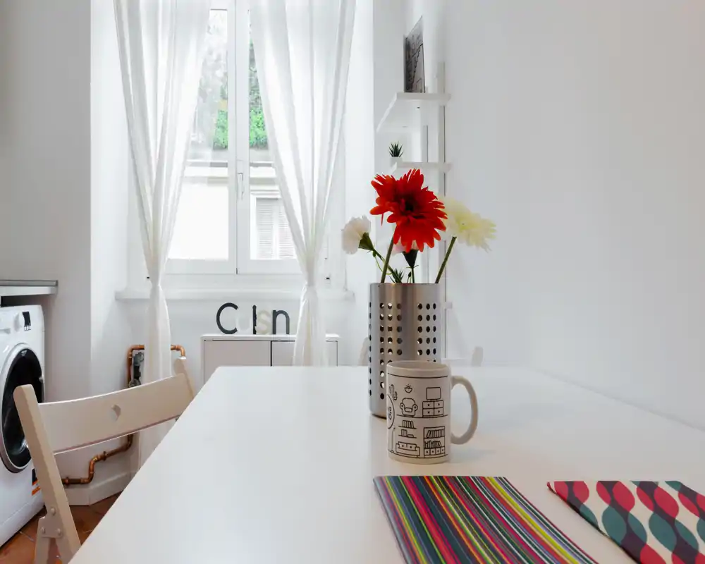 Bright, minimalist kitchen corner with a white table, colorful placemat, mug and fresh flowers on the table; natural light streams through the window and a washing machine is visible in the background.