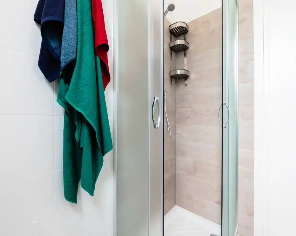 Compact modern bathroom with a glass-enclosed corner shower featuring wood-effect wall panels and a chrome showerhead; colorful towels hang on the adjacent tiled wall.