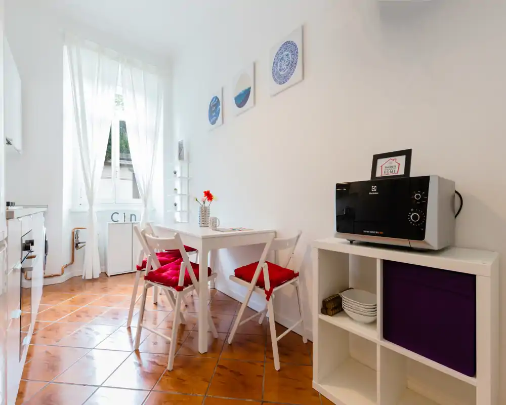 Bright compact kitchen with a small dining nook: white table and chairs with red cushions, tiled floor, microwave on shelving, and a large window providing natural light.