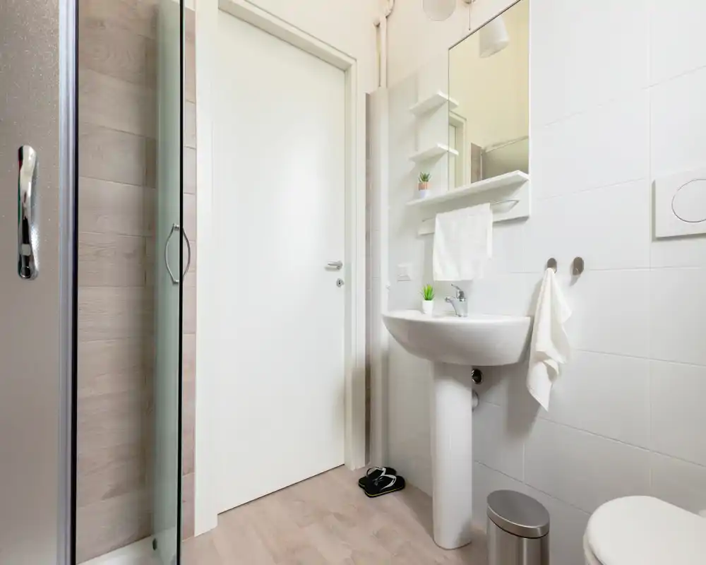 Bright, modern bathroom with a pedestal sink, mirror cabinet and a glass shower door. Clean white tiles and light wood-effect flooring create a fresh, minimal look.