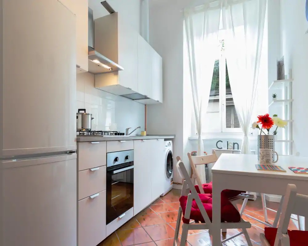 Bright, modern kitchenette with white cabinets, built-in oven, washing machine and a cozy dining corner by a large window.