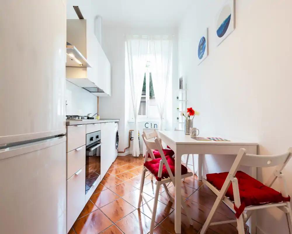 Bright compact kitchen with a white cabinetry line, built-in oven and gas hob, small dining table with red-cushioned chairs and natural light from a tall window.