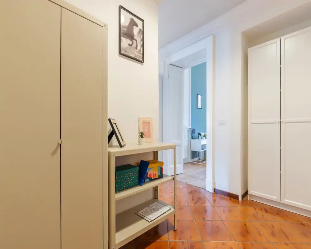 Entrance hallway with tiled floor, storage cabinets and a small shelving unit; bright neutral decor and a visible doorway leading to another room.