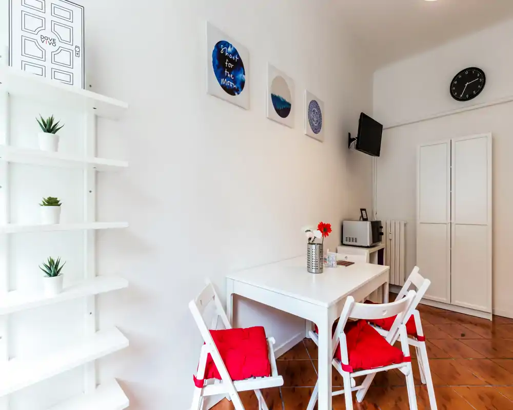 Compact dining area with a white table and four folding chairs accented by red cushions; bright room with simple wall art and small decorative plants.