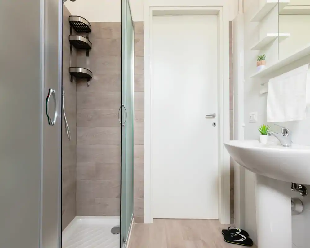 Compact modern bathroom with a glass shower enclosure, white sink and neutral wood-effect tiles — bright and well maintained.