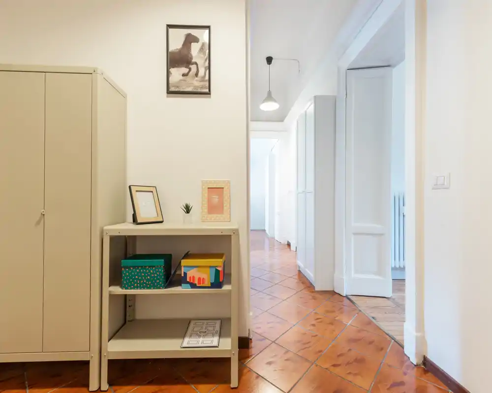Bright, tidy entrance/hallway with tiled floor, a small shelving unit and a wardrobe; clean white walls and a lit corridor leading to other rooms.