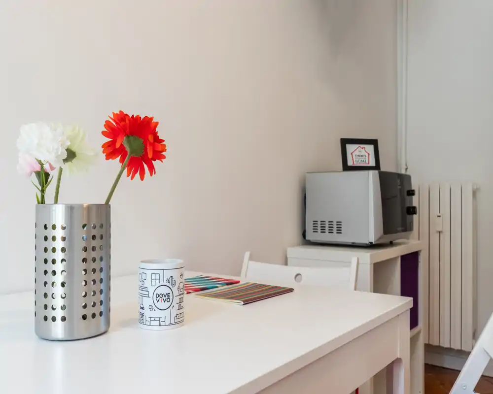 Small, bright kitchen/dining corner with a white table, colorful placemat, decorative flowers and a microwave on a side unit — neat and welcoming for a quick meal.