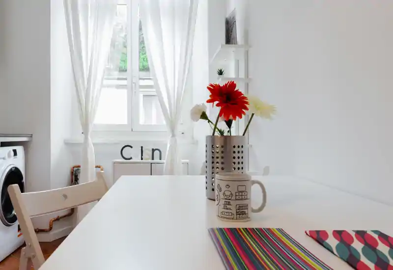 Rincón de cocina luminoso y minimalista con mesa blanca, mantel colorido, taza y flores frescas; luz natural entra por la ventana y se ve una lavadora al fondo.