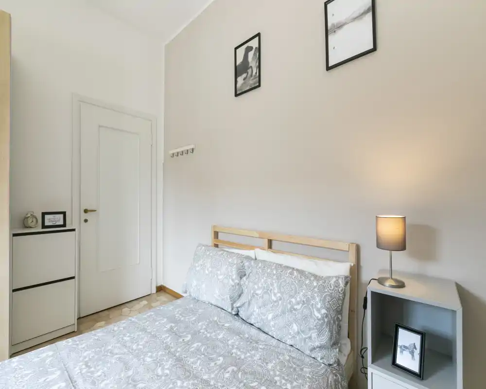 Camera da letto luminosa e ordinata con letto matrimoniale coperto da biancheria a fantasia, comodino con lampada e quadri minimal per un aspetto moderno e rilassante.