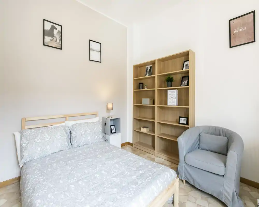 Camera da letto luminosa e semplice con letto matrimoniale, lampada da comodino, libreria in legno e una poltrona comoda — arredamento neutro e pulito, perfetto per un annuncio.