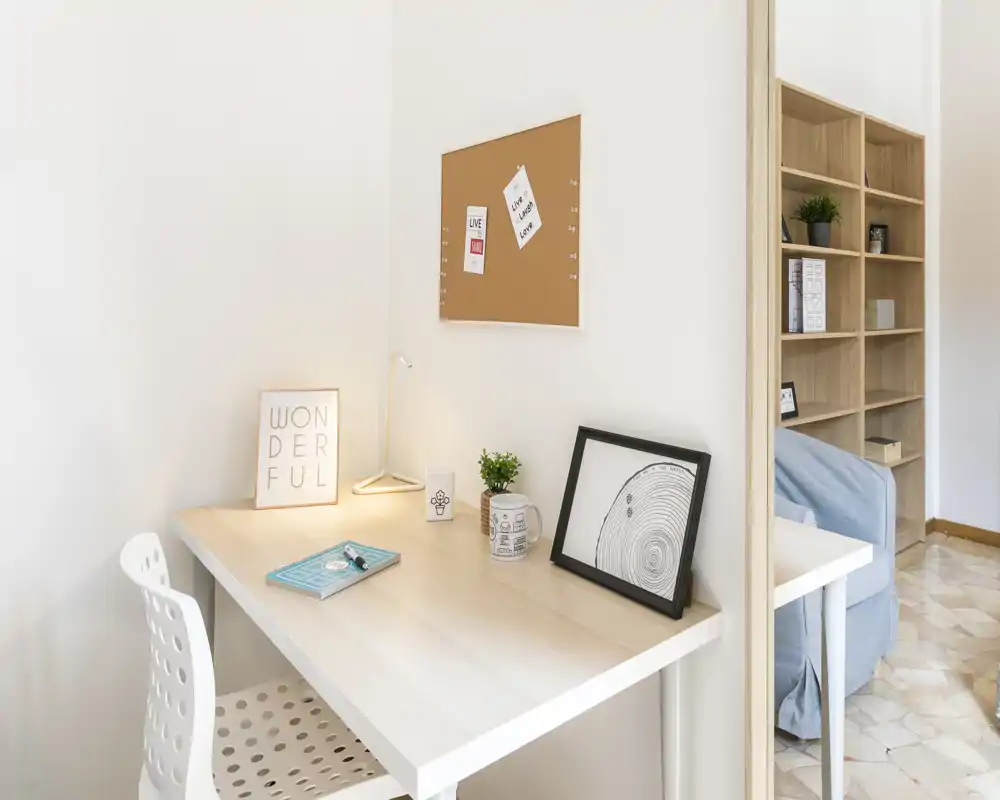 Piccolo spazio di lavoro luminoso contro una parete bianca con scrivania in legno chiaro, lampada, sedia e oggetti decorativi; libreria e divano sono visibili nell'area soggiorno adiacente.