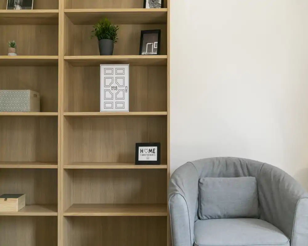 Angolo soggiorno elegante con una poltrona grigia morbida accanto a scaffalature in legno con pochi elementi decorativi, che crea un'atmosfera calma e accogliente.
